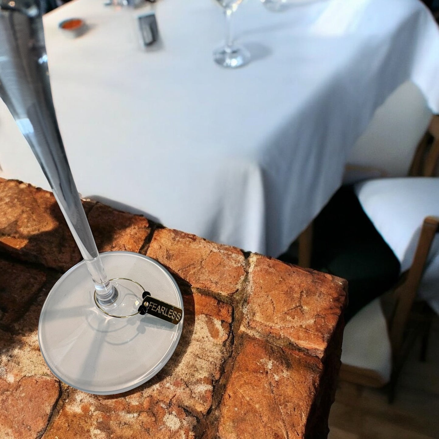 Wine glass charm with a 'FEARLESS' label on a stone surface, blurred restaurant setting in the background