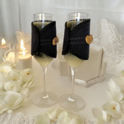 Two champagne glasses with black sleeves and gold buttons on a decorative table with candles and flowers.