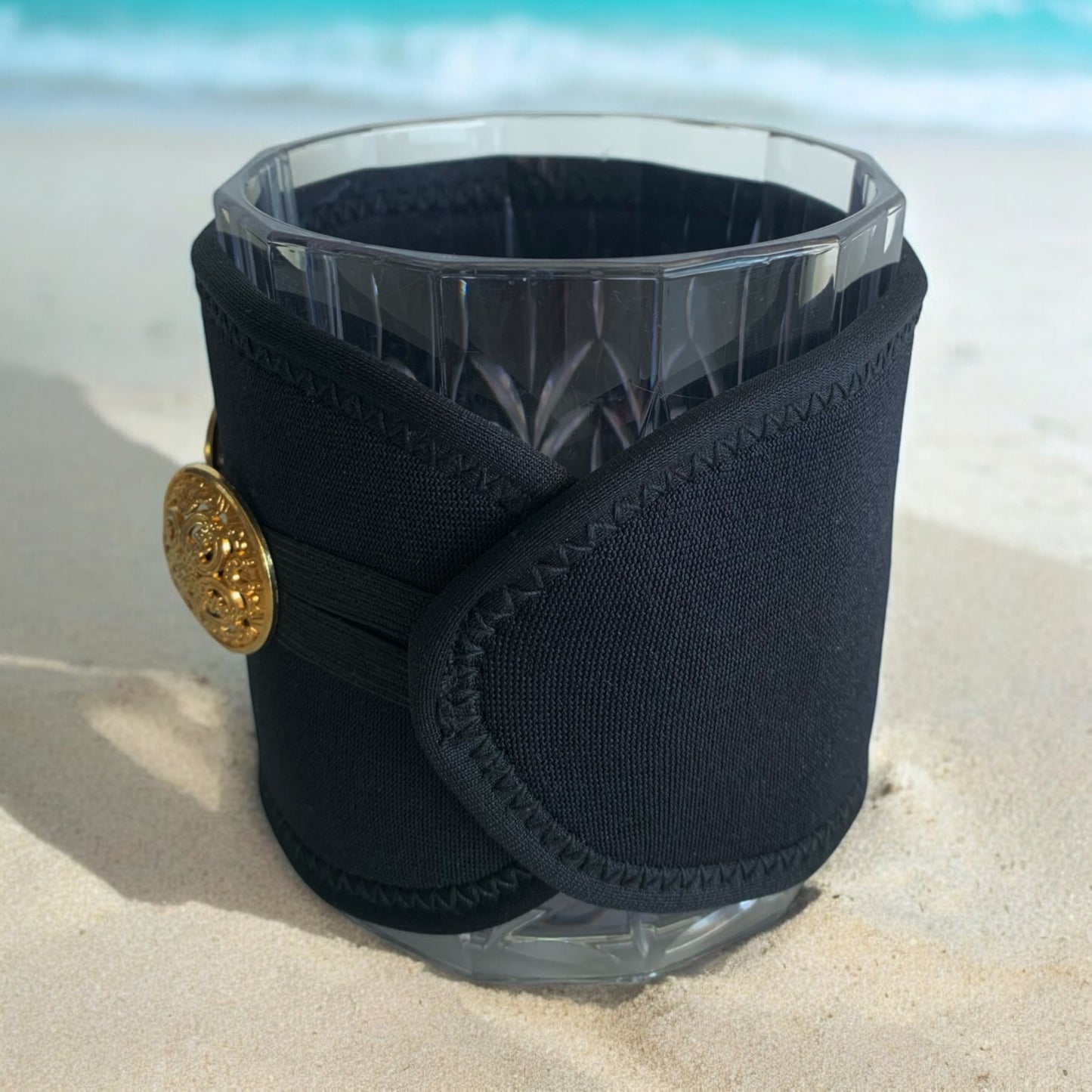 Black neoprene cup holder with gold button on a clear glass against a beach background