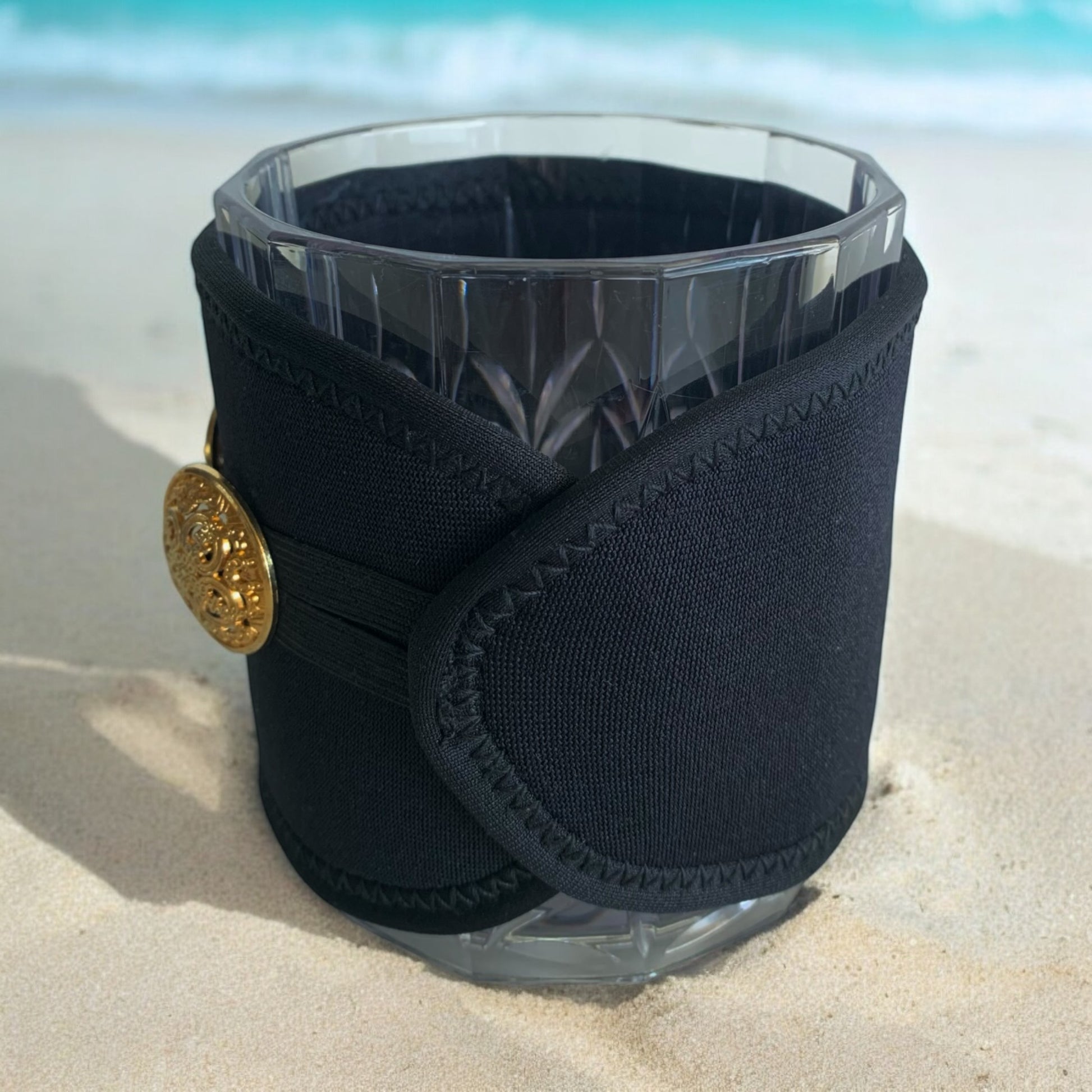 Black neoprene cup holder with gold button on a clear glass against a beach background