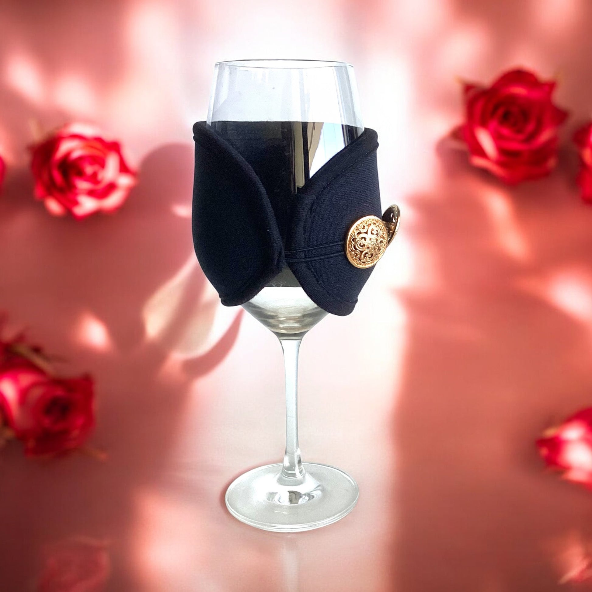 A black insulated wine glass wrap with an adjustable band and a decorative button, displayed on a wine glass with a pink and rose petal background.